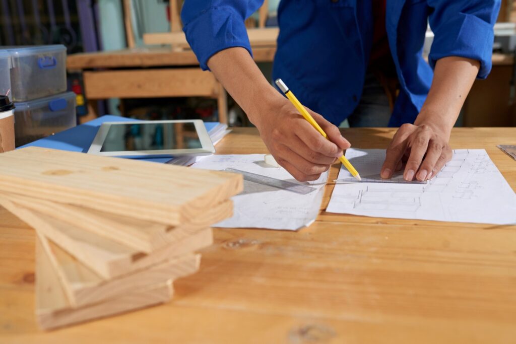 Person in a blue shirt drawing architectural plans with a yellow pencil and ruler on a wooden table; blueprint sheets and a tablet nearby.